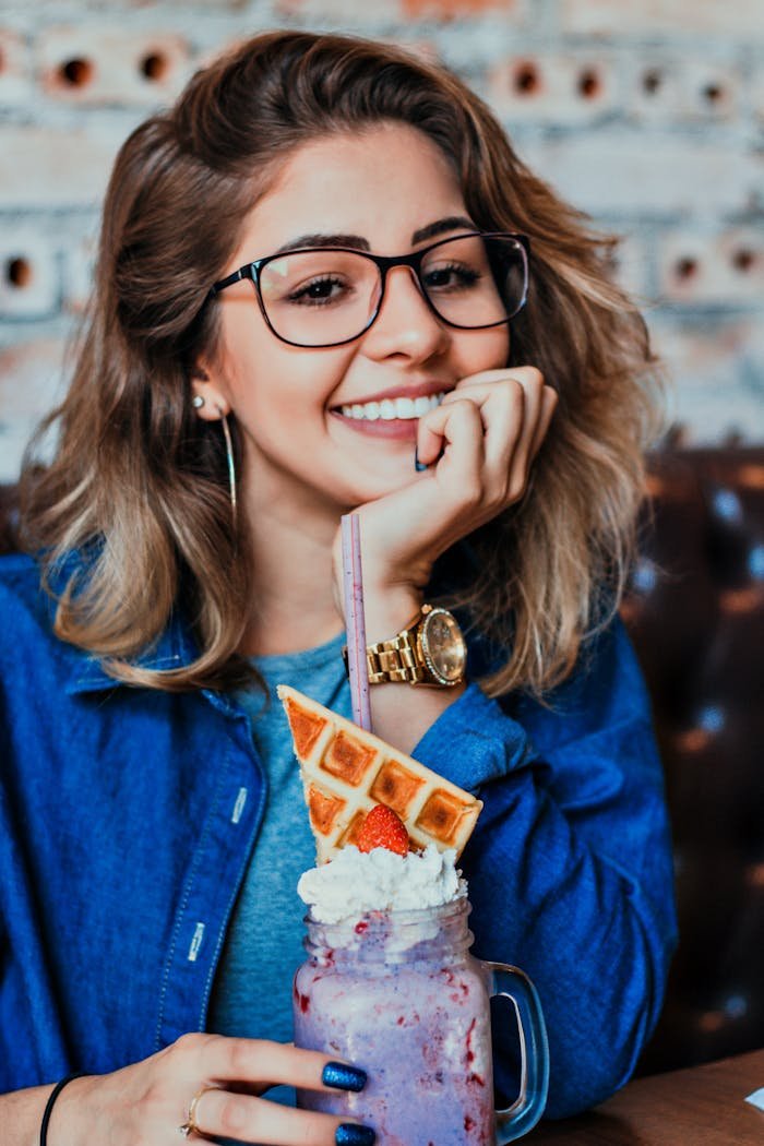 A smiling woman in glasses enjoying a colorful beverage indoors, featuring waffles and whipped cream.